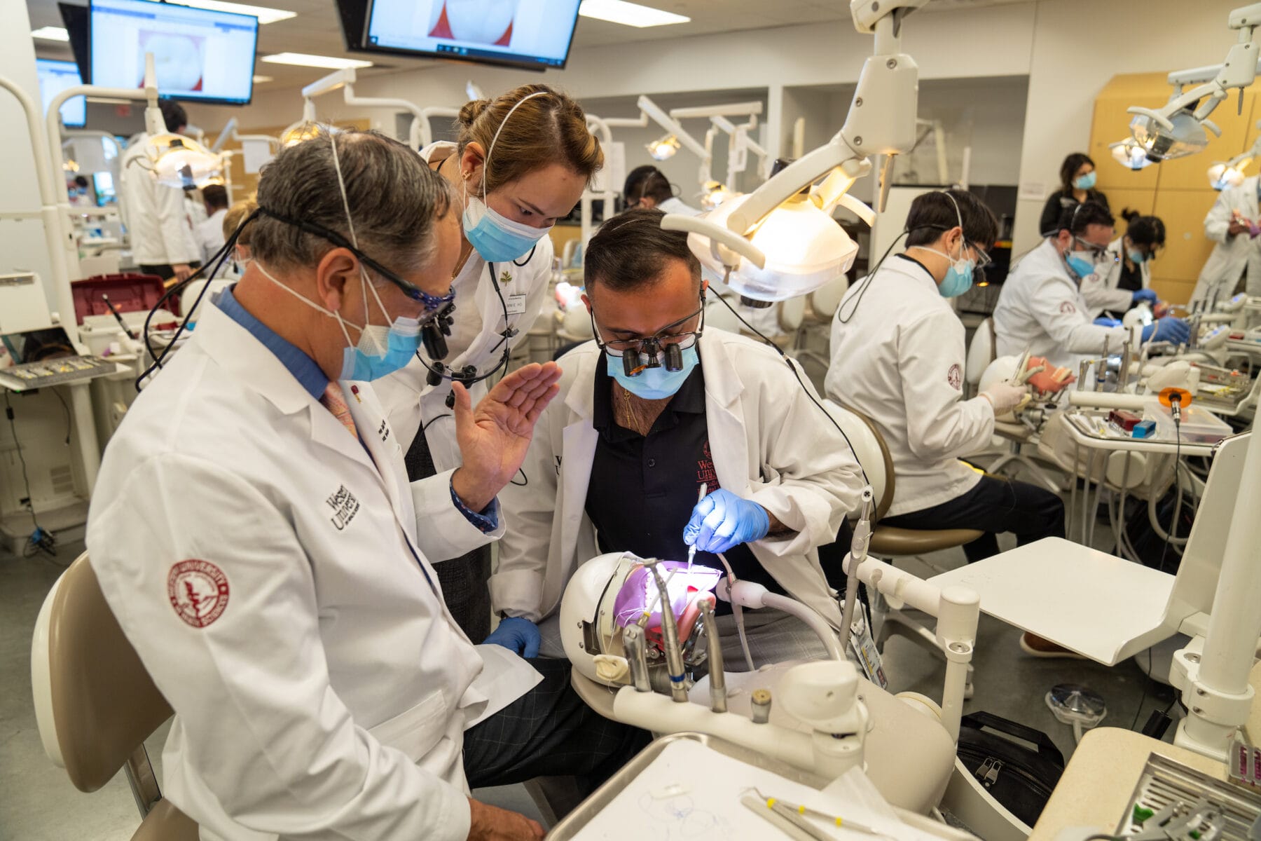 Dental students and instructors in white coats and masks work on dental mannequins in a clinical classroom setting, with dental tools and monitors visible.