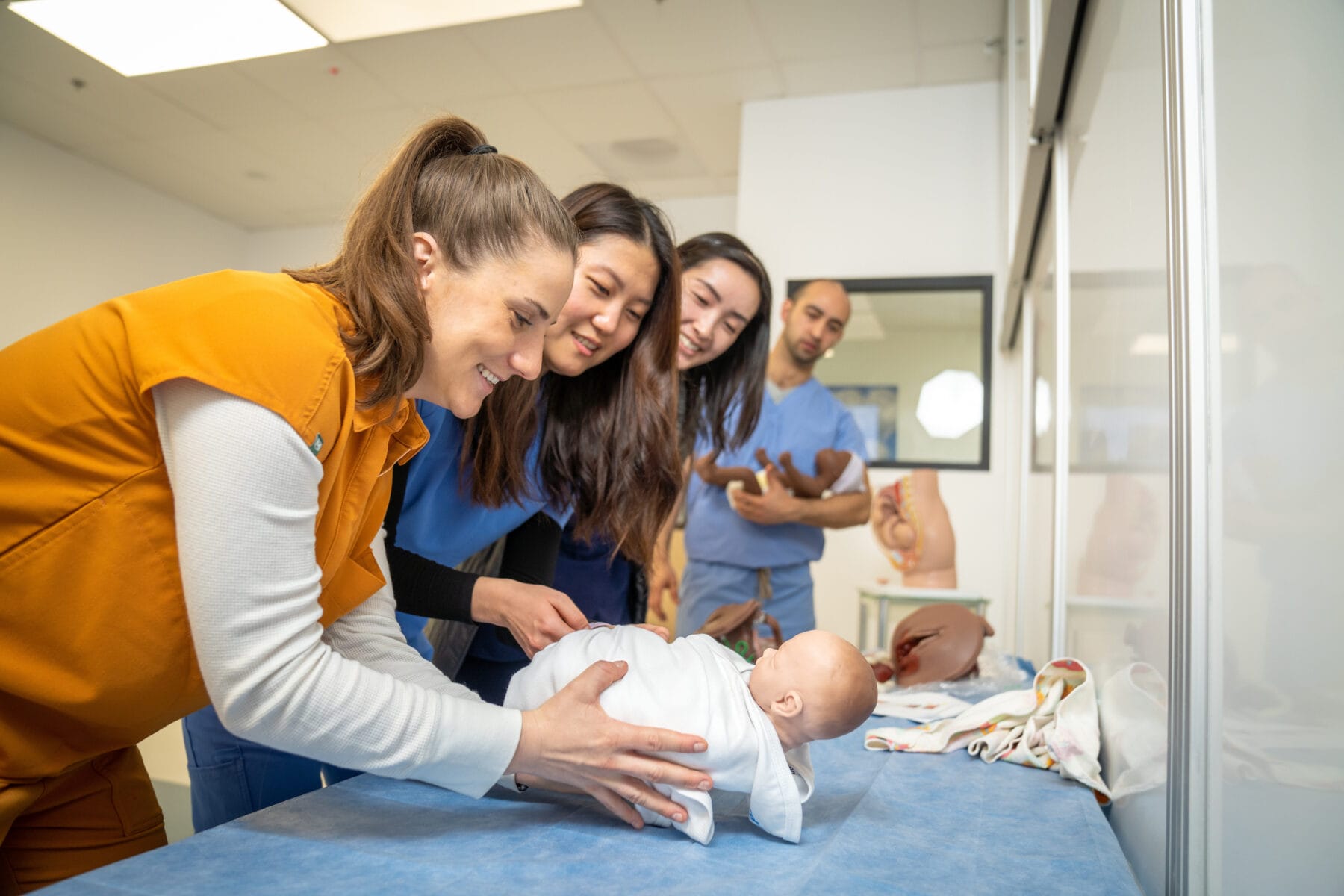 Four medical students practice swaddling a baby mannequin on a table in a clinical classroom setting.