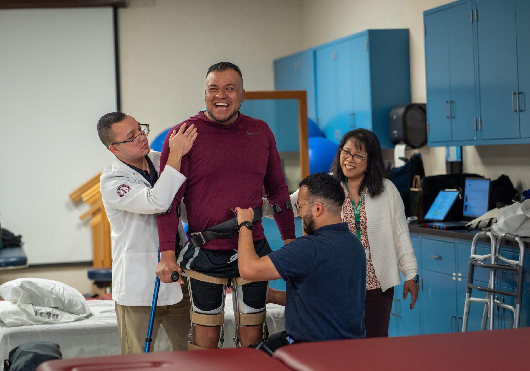 A man in a maroon shirt uses leg braces and a walker while assisted by three people in a rehabilitation clinic.