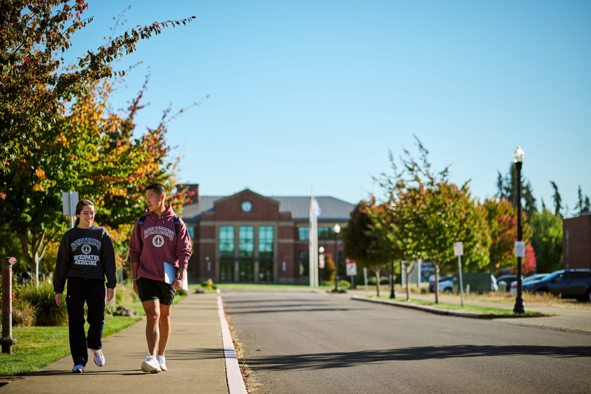 Two people walk on a sidewalk beside a tree-lined street near a brick building on a clear day.