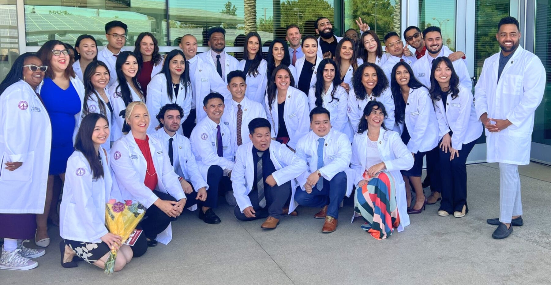 A group of students in white coats pose for a photo outside a building; some are standing, others are kneeling or crouching, and one person holds a bouquet of flowers.