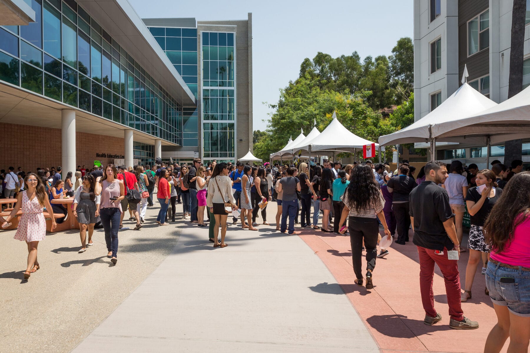 A large group of people gathers outdoors between modern buildings and white tents on a sunny day, some standing in lines, while others walk or talk.