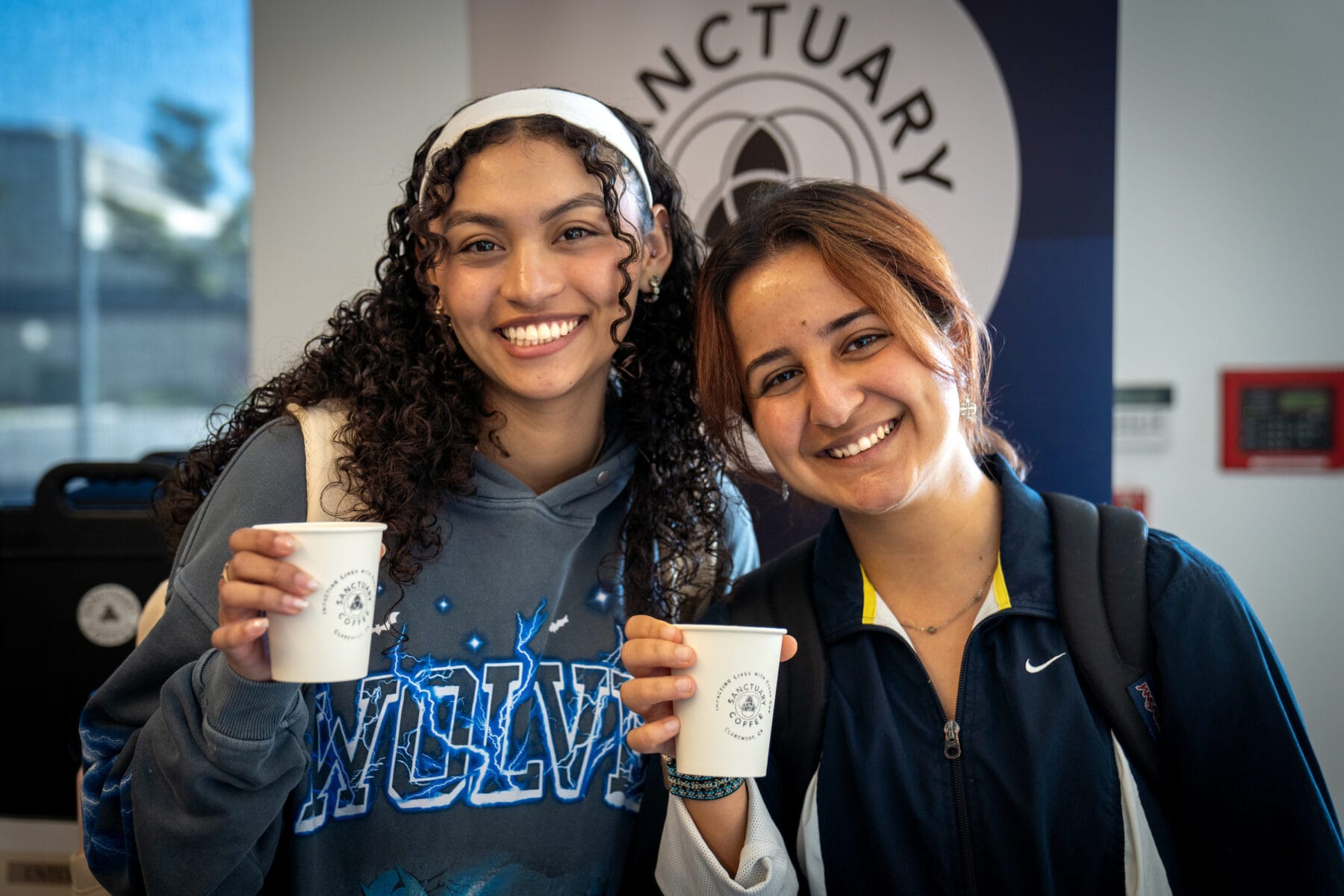 Two young women smile and hold paper cups in front of a "Sanctuary" sign. One wears a "Wolves" hoodie, the other a dark jacket with a backpack.