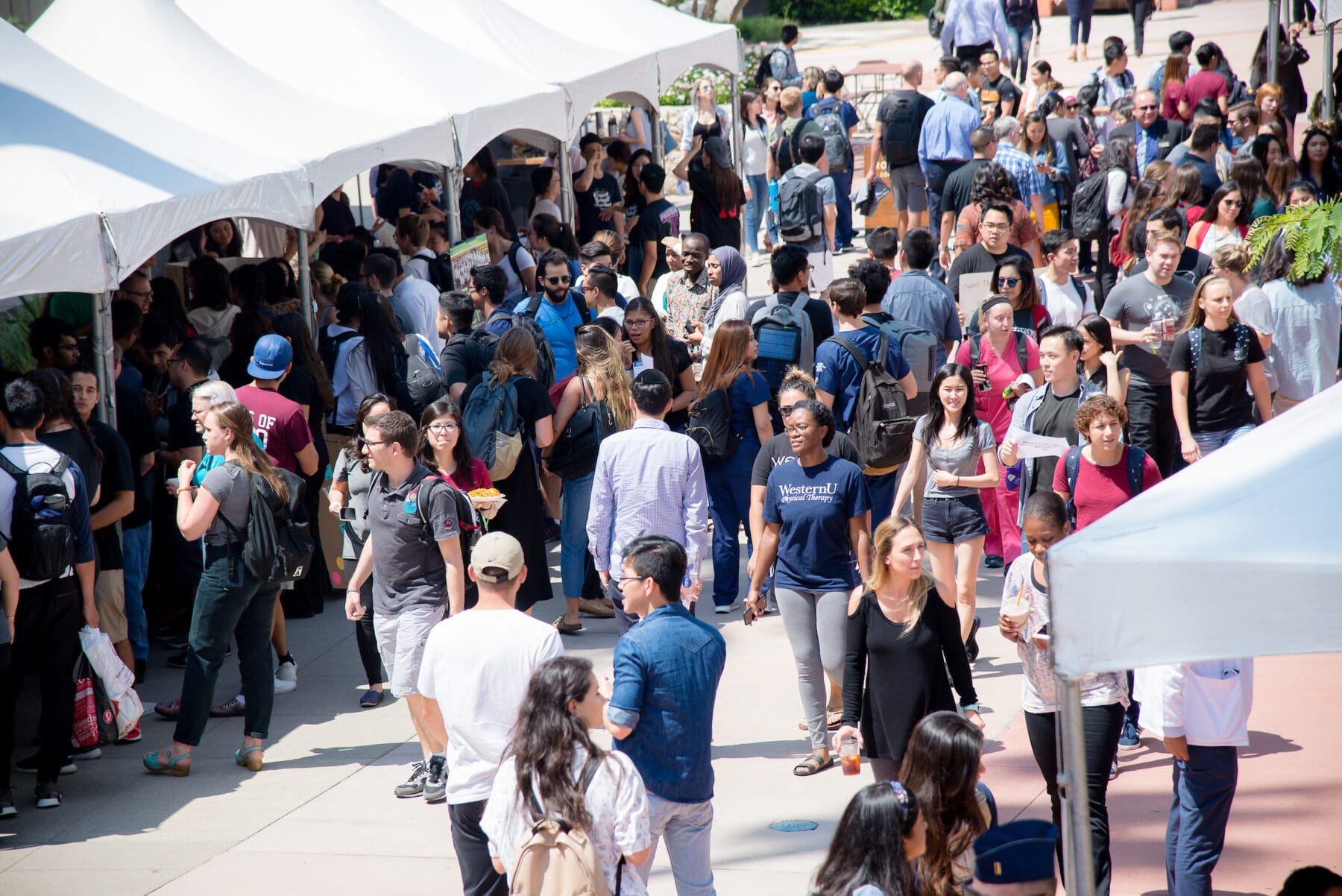 A large crowd of people, mostly young adults, walk and gather under tents outdoors on a sunny day, suggesting a busy campus or event setting.