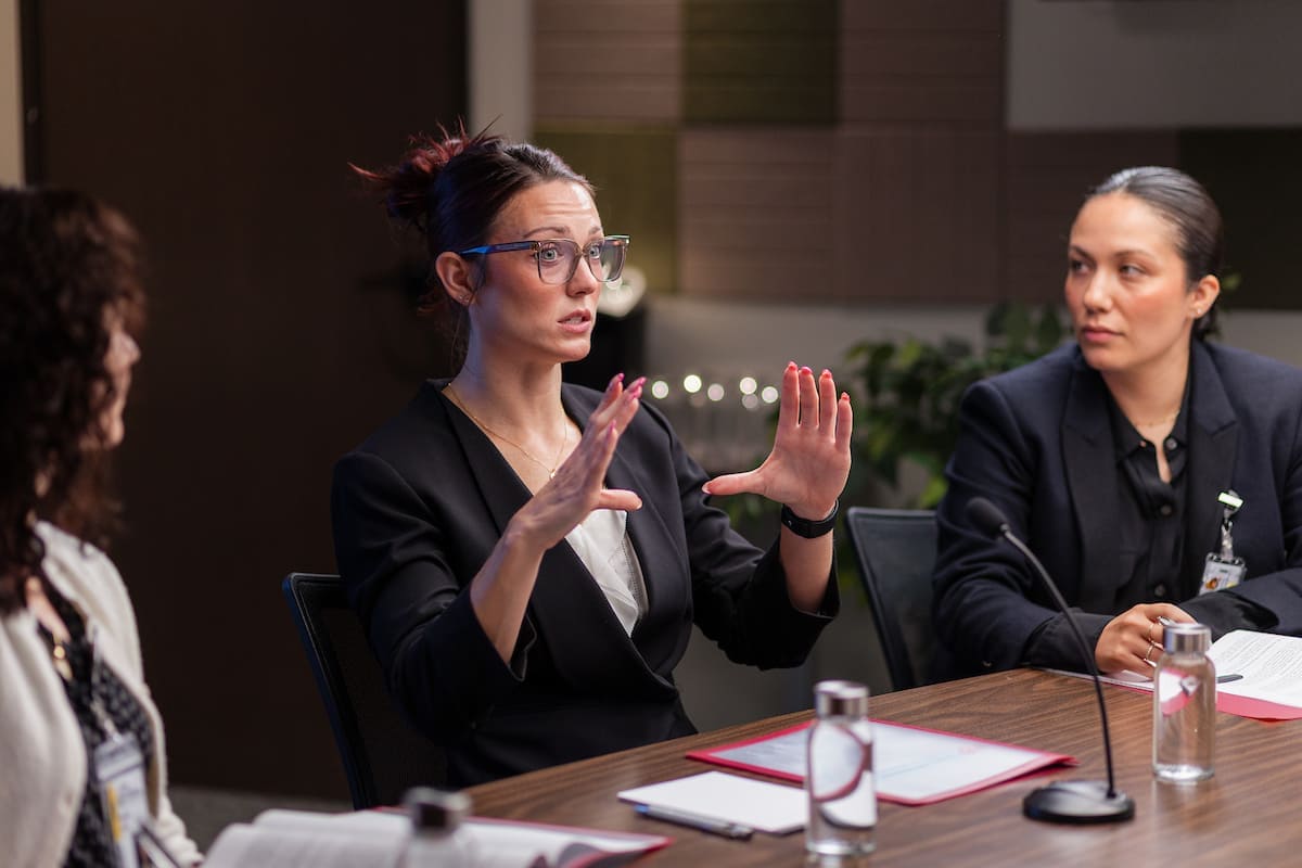 A woman in a blazer gestures while speaking to colleagues during a meeting at a conference table.