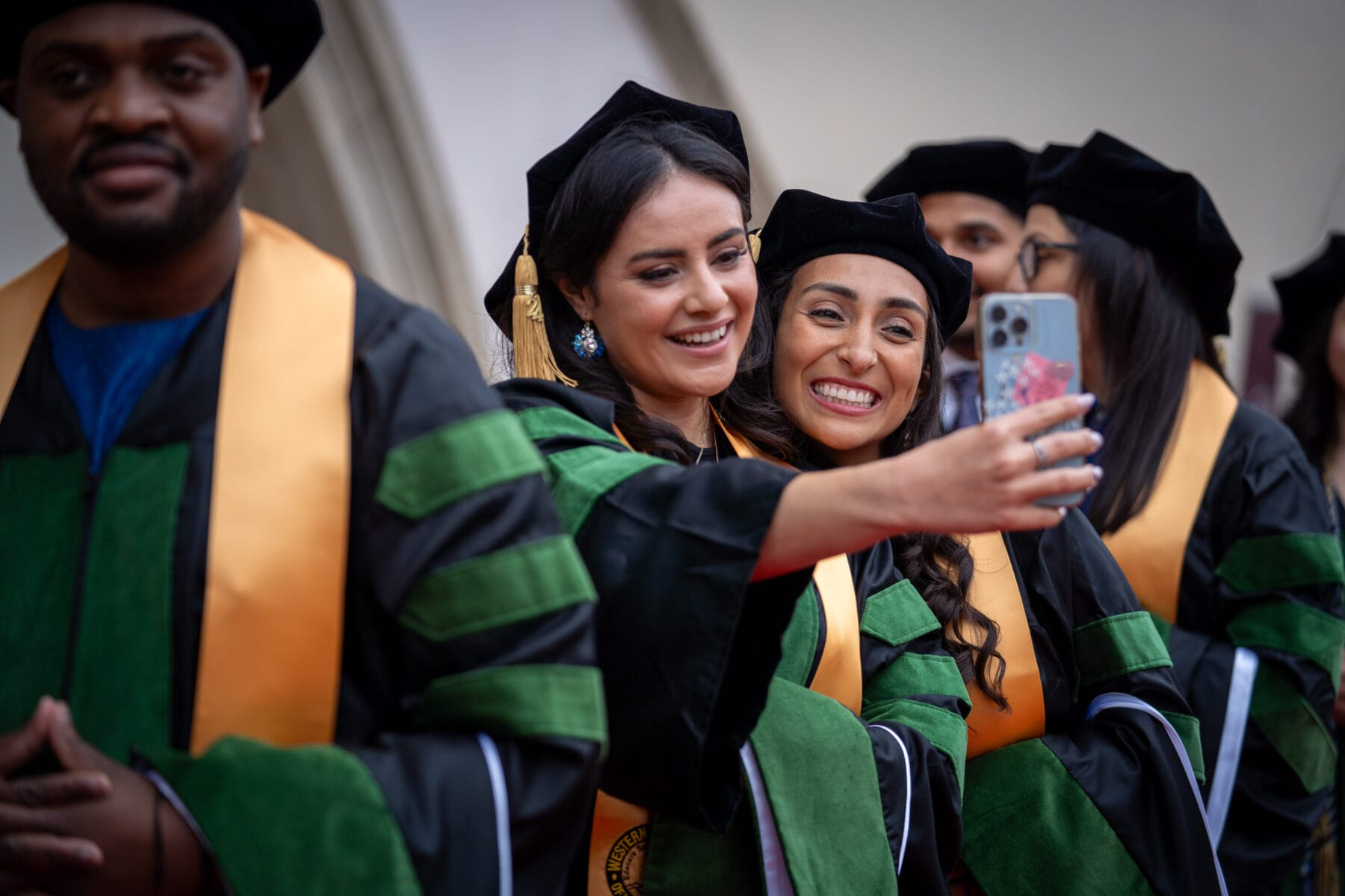 Two women. In graduation robes. Hugging, smiling. One is holding a cell phone in her hand and taking a photo.