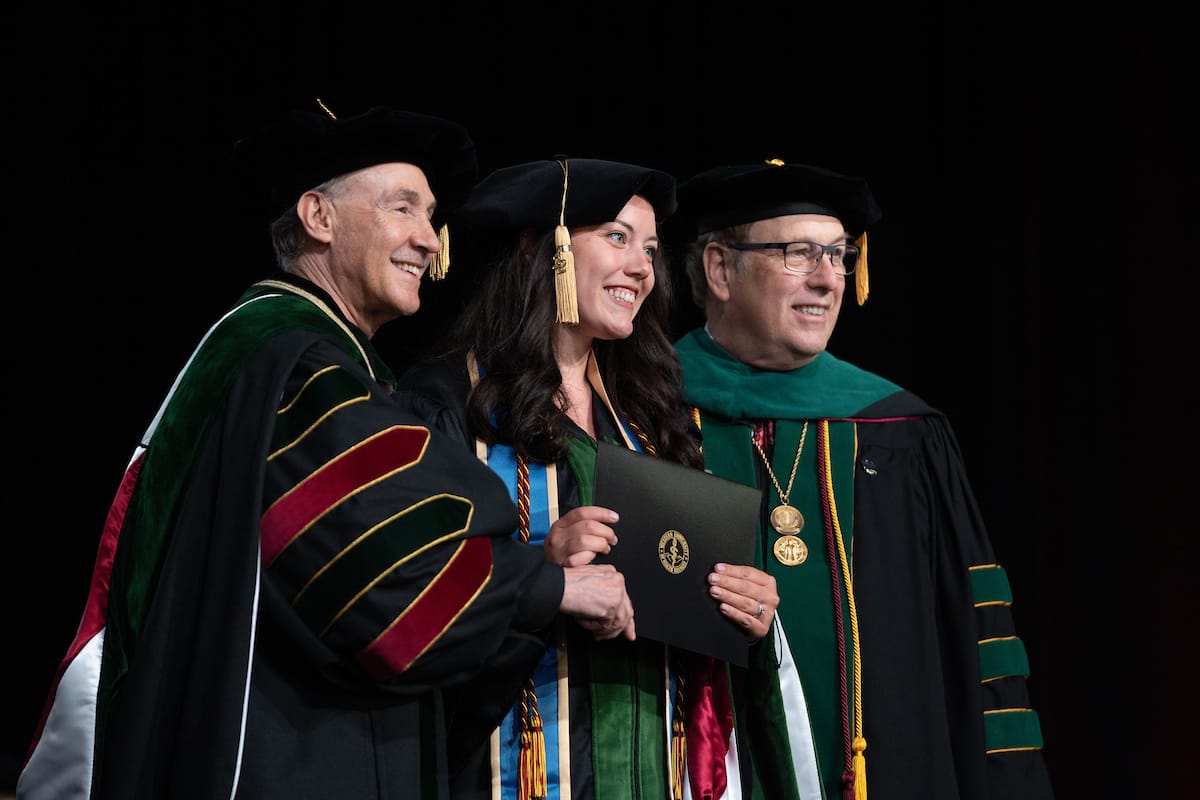 Three people in graduation attire smile and pose for a photo. The middle person holds a diploma while the others stand on either side.
