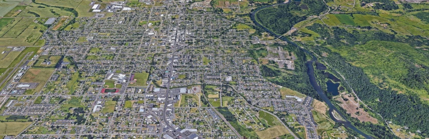 Aerial view of a town with a river flowing through it, surrounded by fields and forests. Roads and buildings form a grid-like pattern.