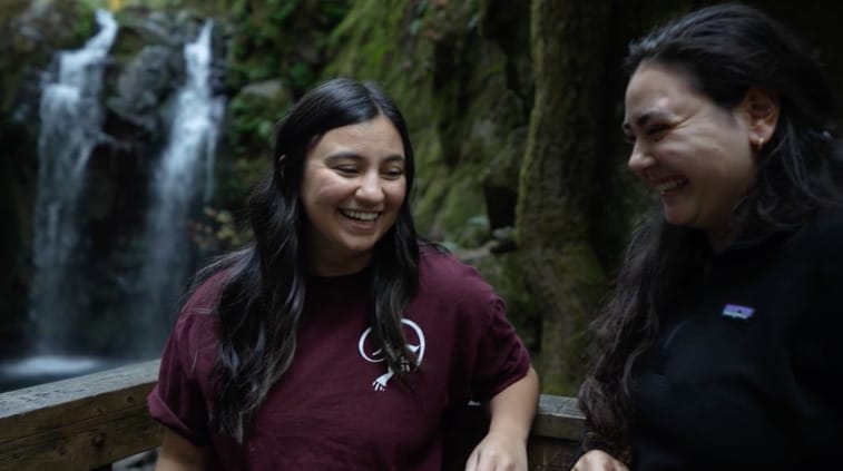 Two women stand on a wooden railing in front of a waterfall, smiling and laughing. The background is green and mossy.