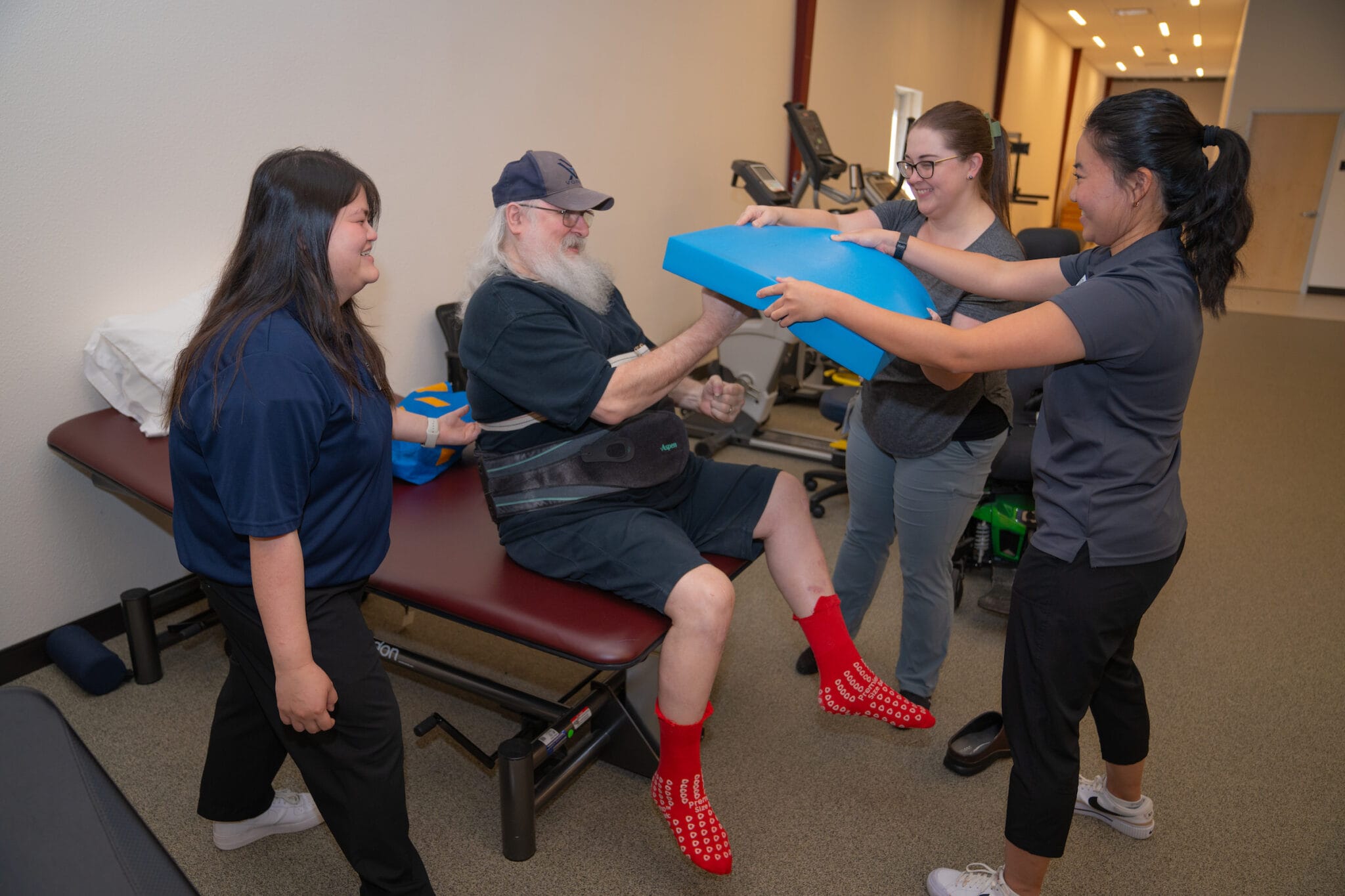 An older man sits on a therapy table, assisted by three women during a rehabilitation exercise using a blue foam pad.
