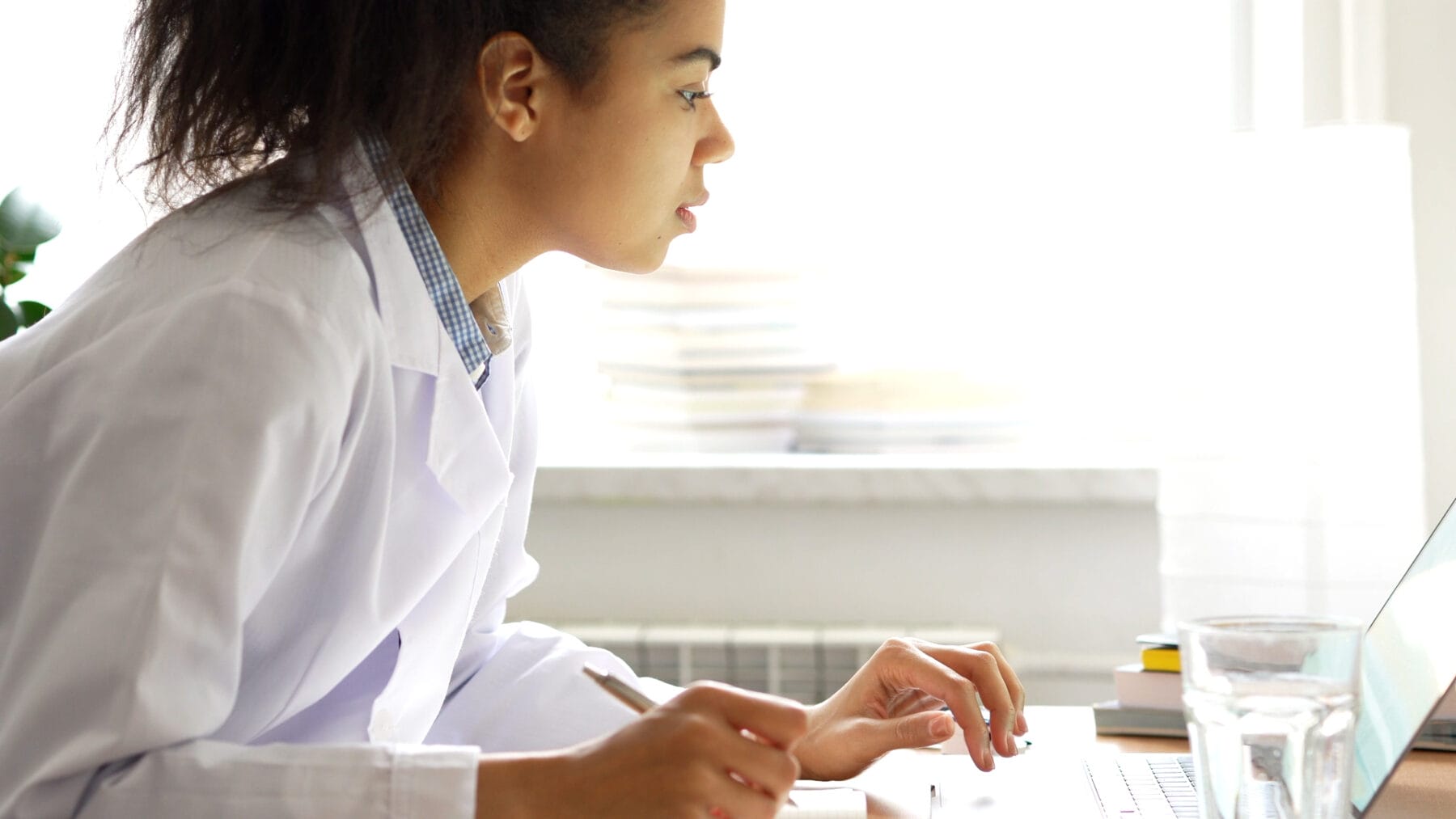 A person wearing a white lab coat sits at a desk, focused on a laptop screen with a pen in hand. There are stacks of books and a glass of water on the desk.
