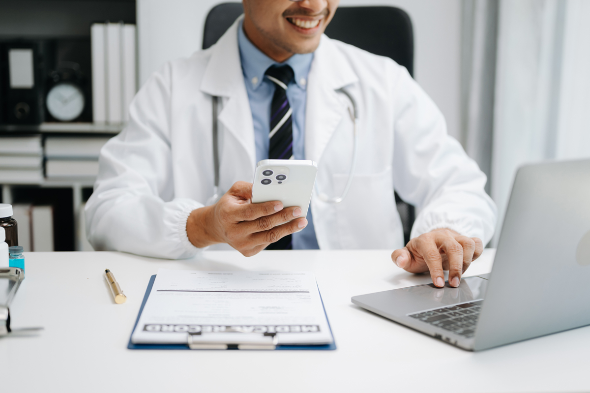 A doctor holding a cell phone in front of a laptop.