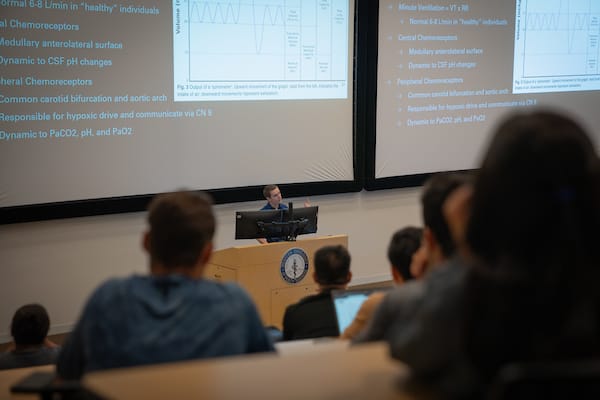 A lecturer stands at a podium presenting slides with medical or scientific data to students in a university lecture hall.