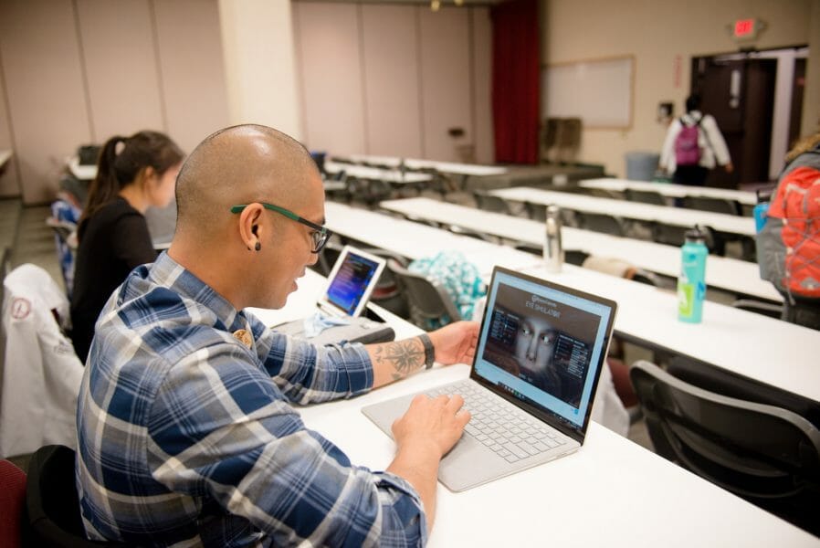 A person in a plaid shirt and glasses uses a laptop in a classroom, with other students and laptops visible in the background.