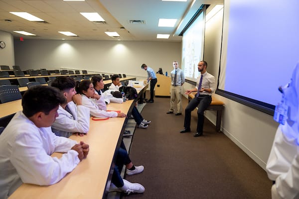 A group of students in lab coats sit at desks while two instructors speak at the front of a classroom with a projection screen.