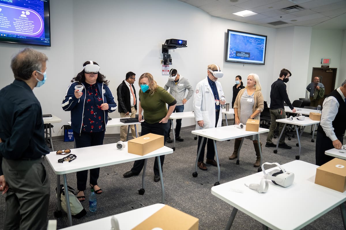 People in a classroom are using virtual reality headsets and controllers. Some individuals are standing while others are seated at tables with VR equipment. Everyone is wearing masks.