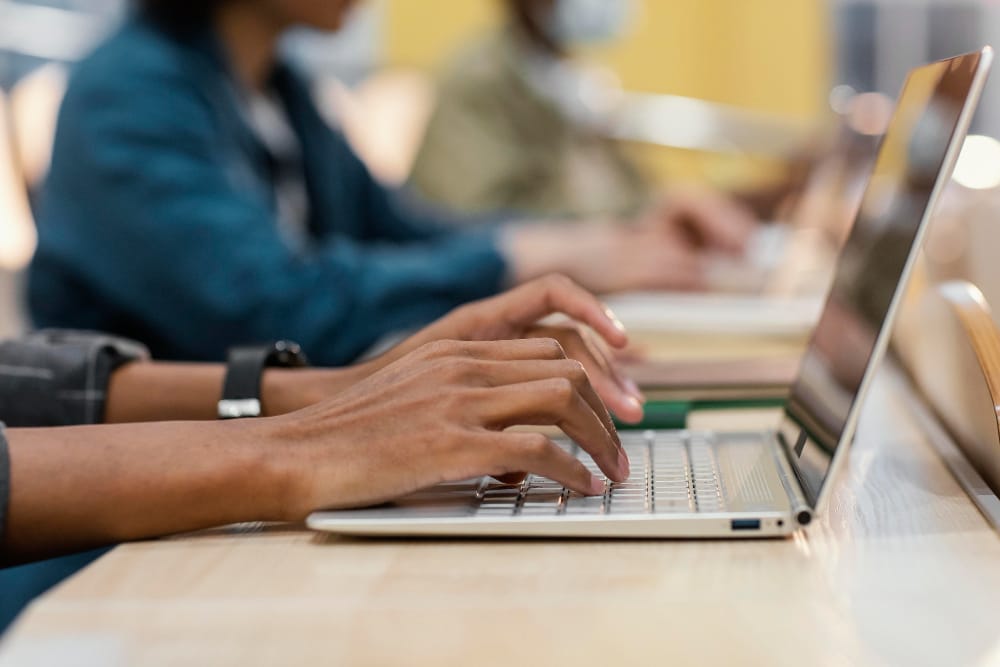 Close-up of hands typing on a laptop keyboard, with other people using laptops in the background at a shared workspace or classroom.