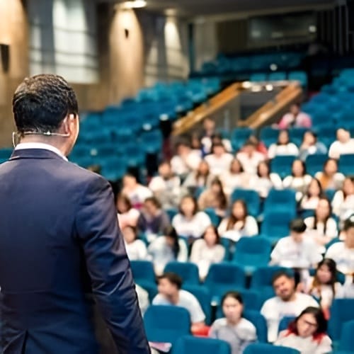 A person in a suit speaks to an audience seated in a large auditorium.