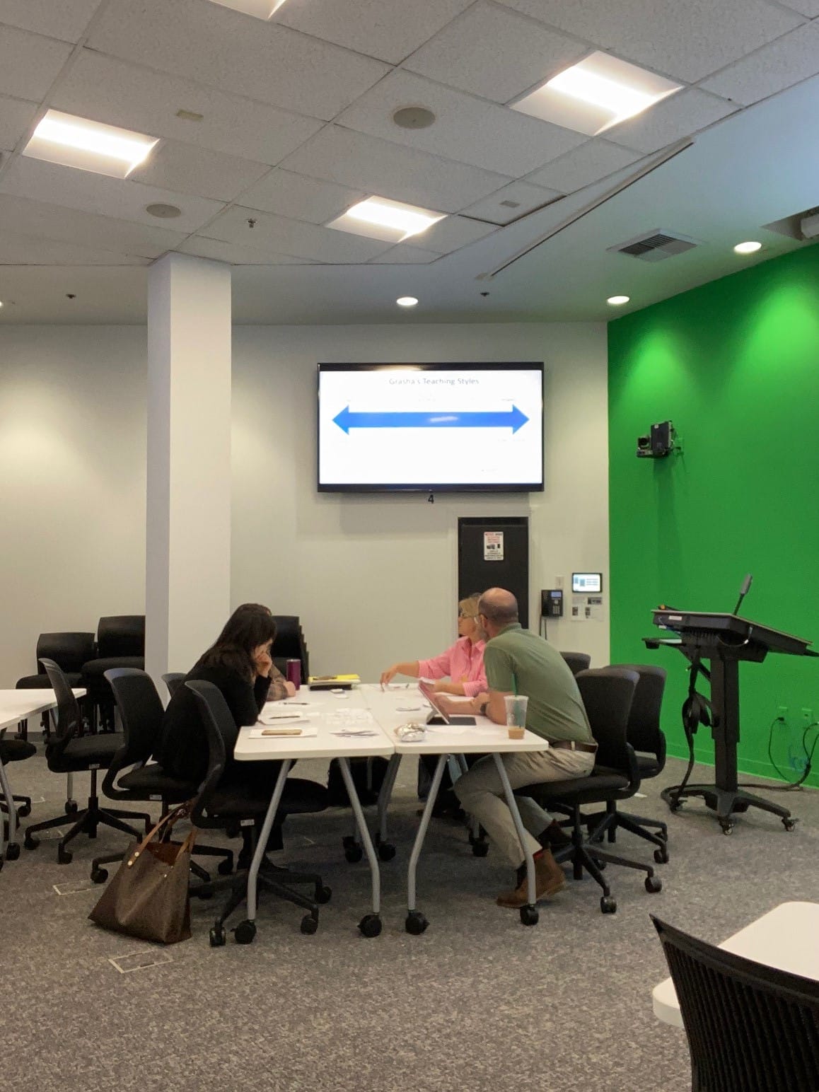Two people sit at a table engaged in discussion in a conference room. A presentation slide is displayed on a screen behind them. The room features a green accent wall and several chairs and tables.