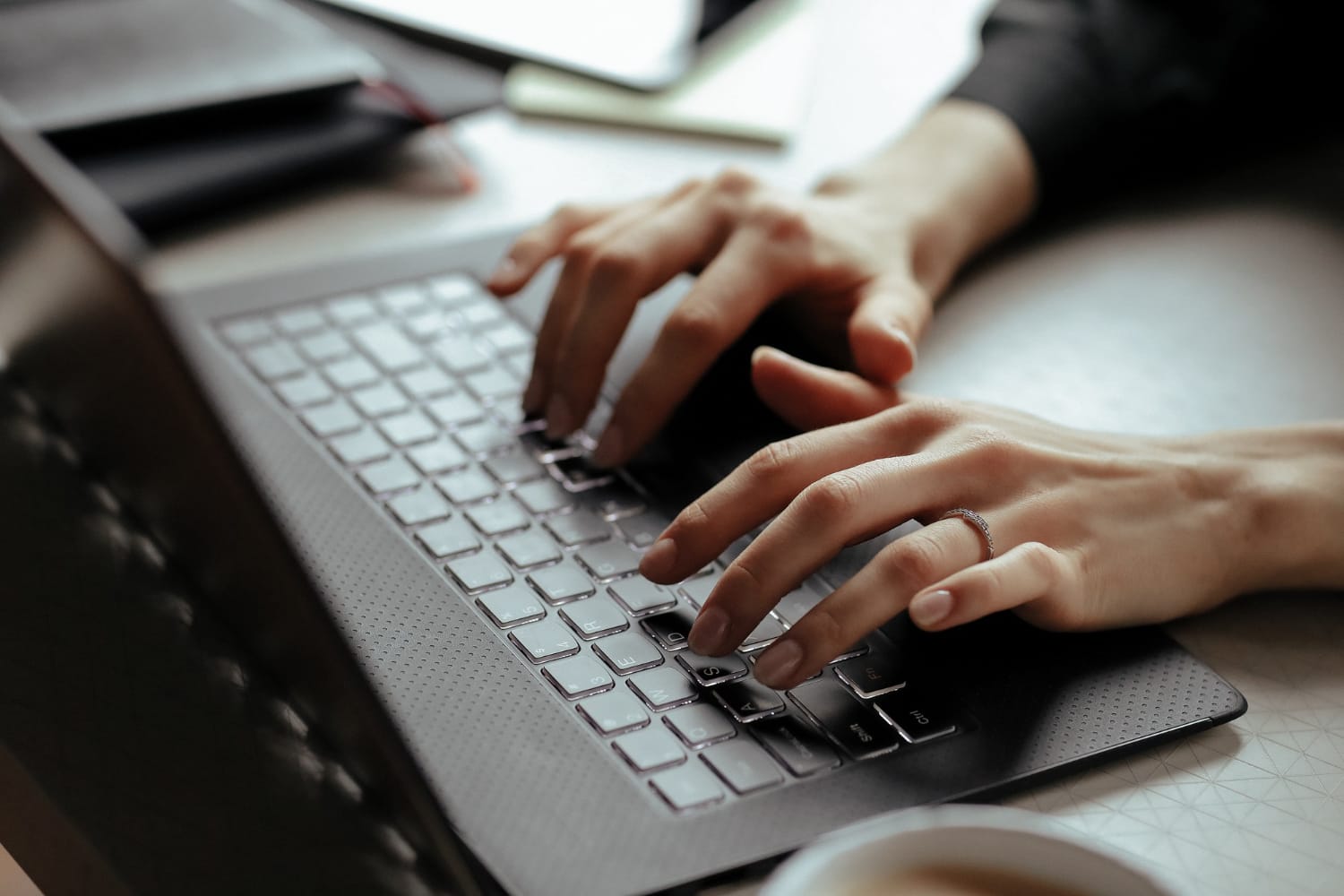Close-up of hands typing on a laptop keyboard, with a desk and some office items blurred in the background.