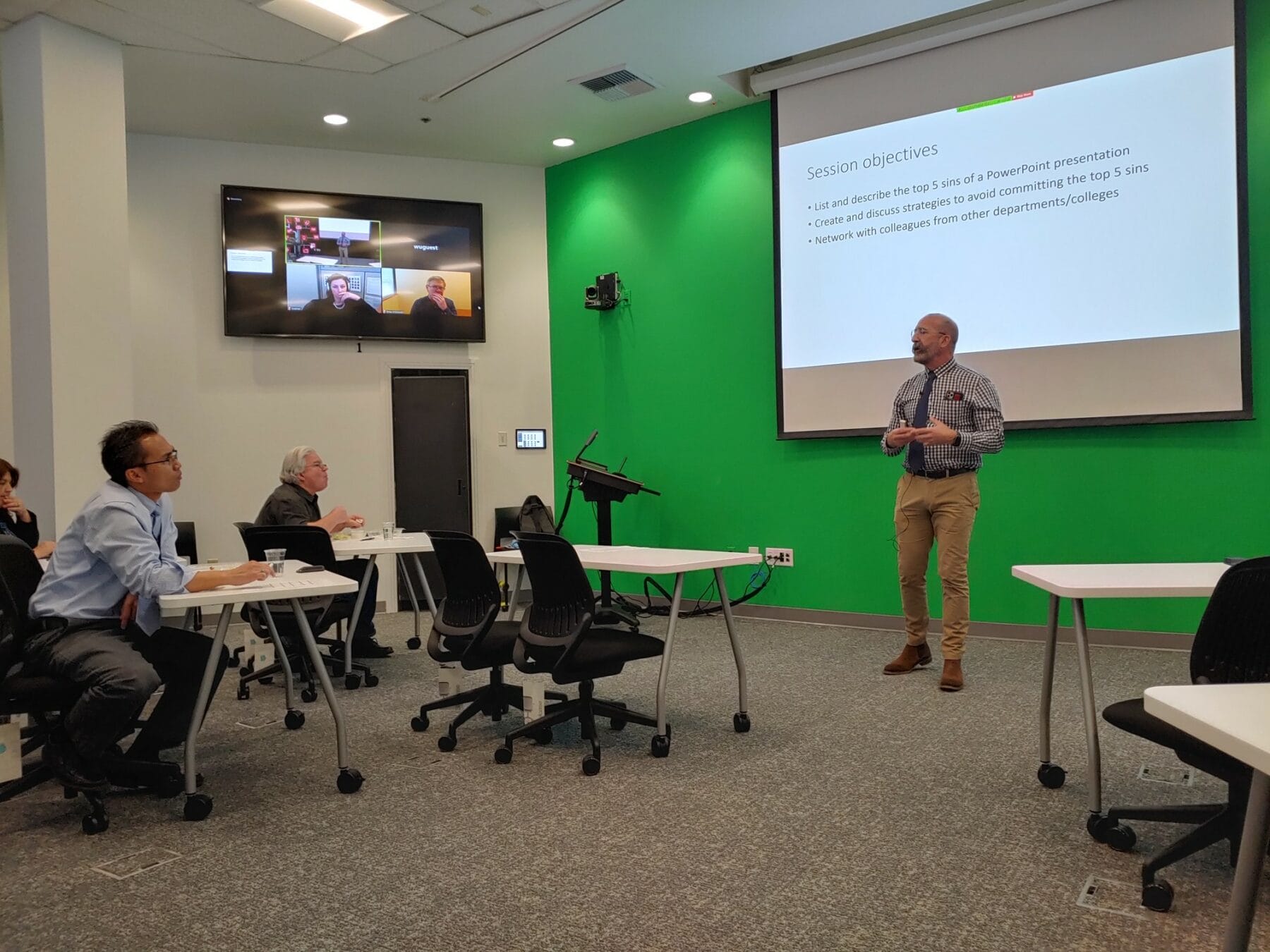 A person is standing and presenting in a conference room with session objectives displayed on a screen. Other attendees are seated and one interacts with a remote participant via video call.