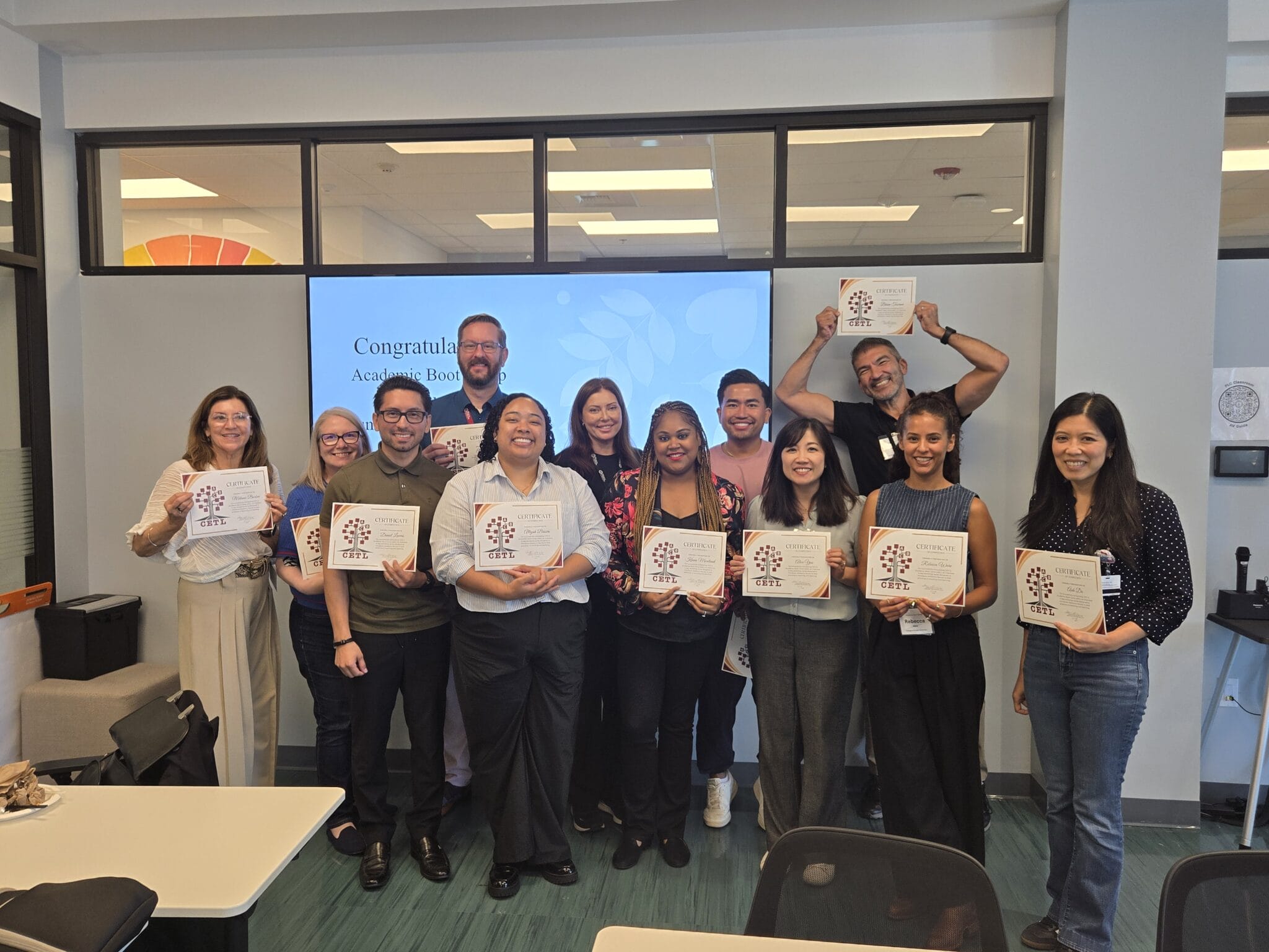 Twelve people stand indoors, smiling and holding certificates, posing for a group photo in front of a screen displaying a congratulatory message.