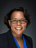Portrait of a smiling woman with glasses, wearing a blue blouse and a dark blazer.