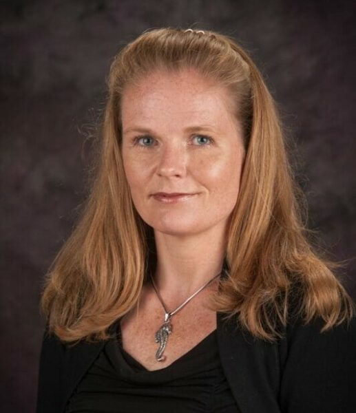 A professional portrait of a woman with long blonde hair, wearing a black top and a silver pendant, against a dark mottled background.