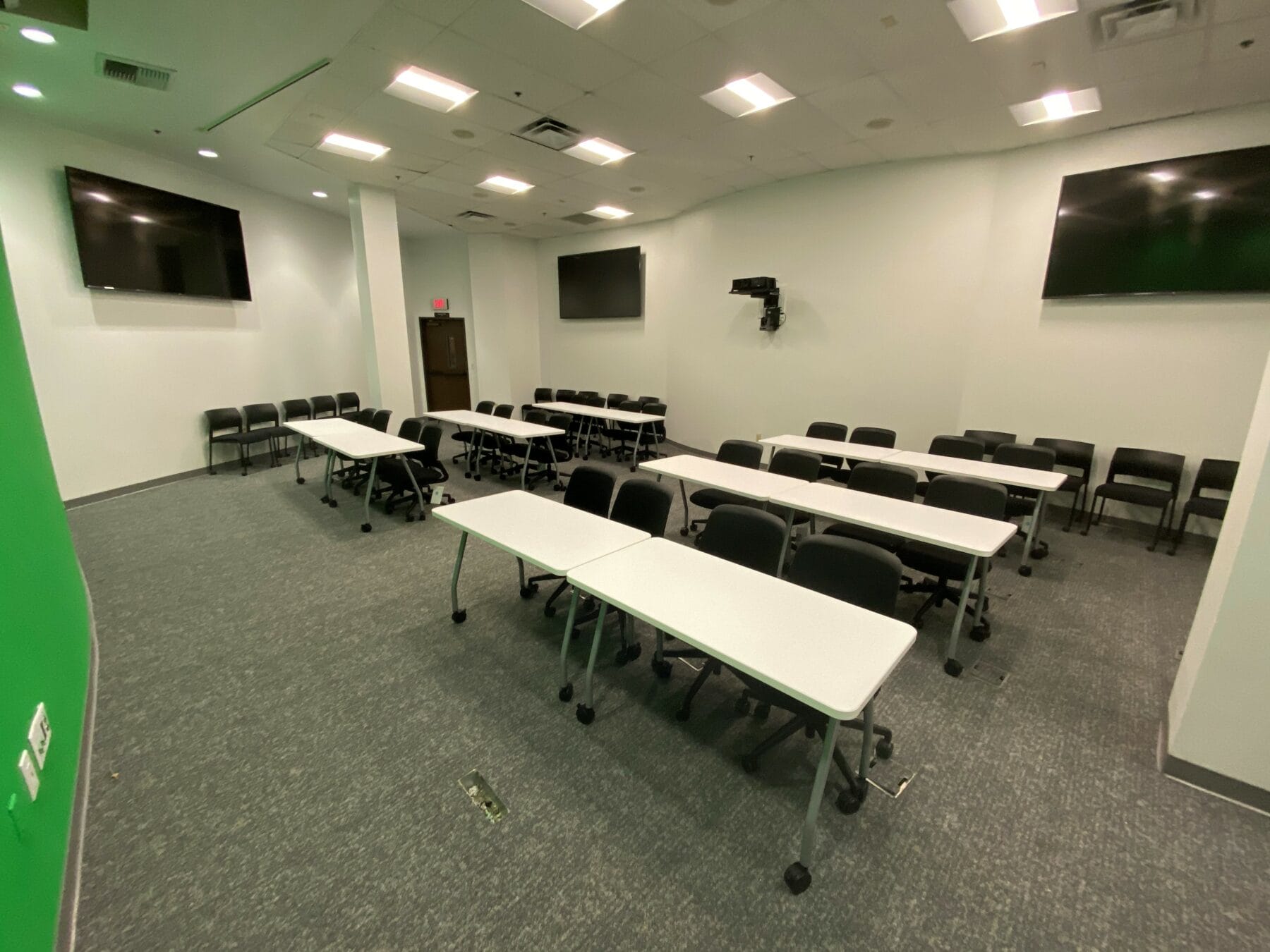 A conference room with several tables and chairs arranged facing a wall with three mounted flat-screen monitors, well-lit with fluorescent lights, and a green screen on the left side.