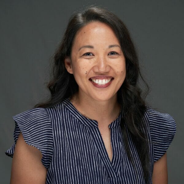 A smiling asian woman with long dark hair, wearing a striped sleeveless top, against a gray background.