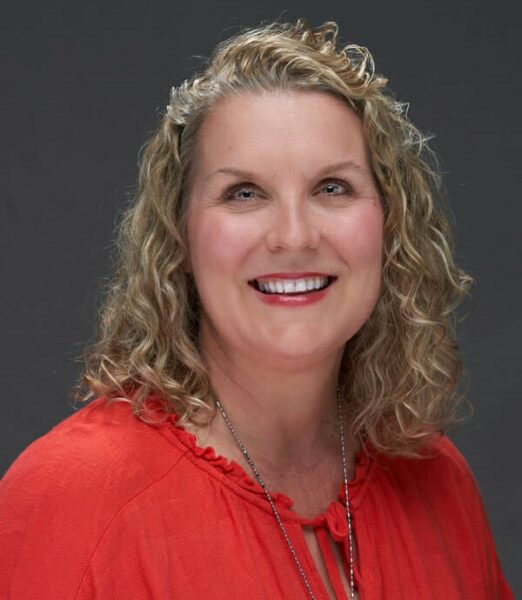 Middle-aged woman with curly hair, wearing a red blouse and a necklace, smiling against a gray background.