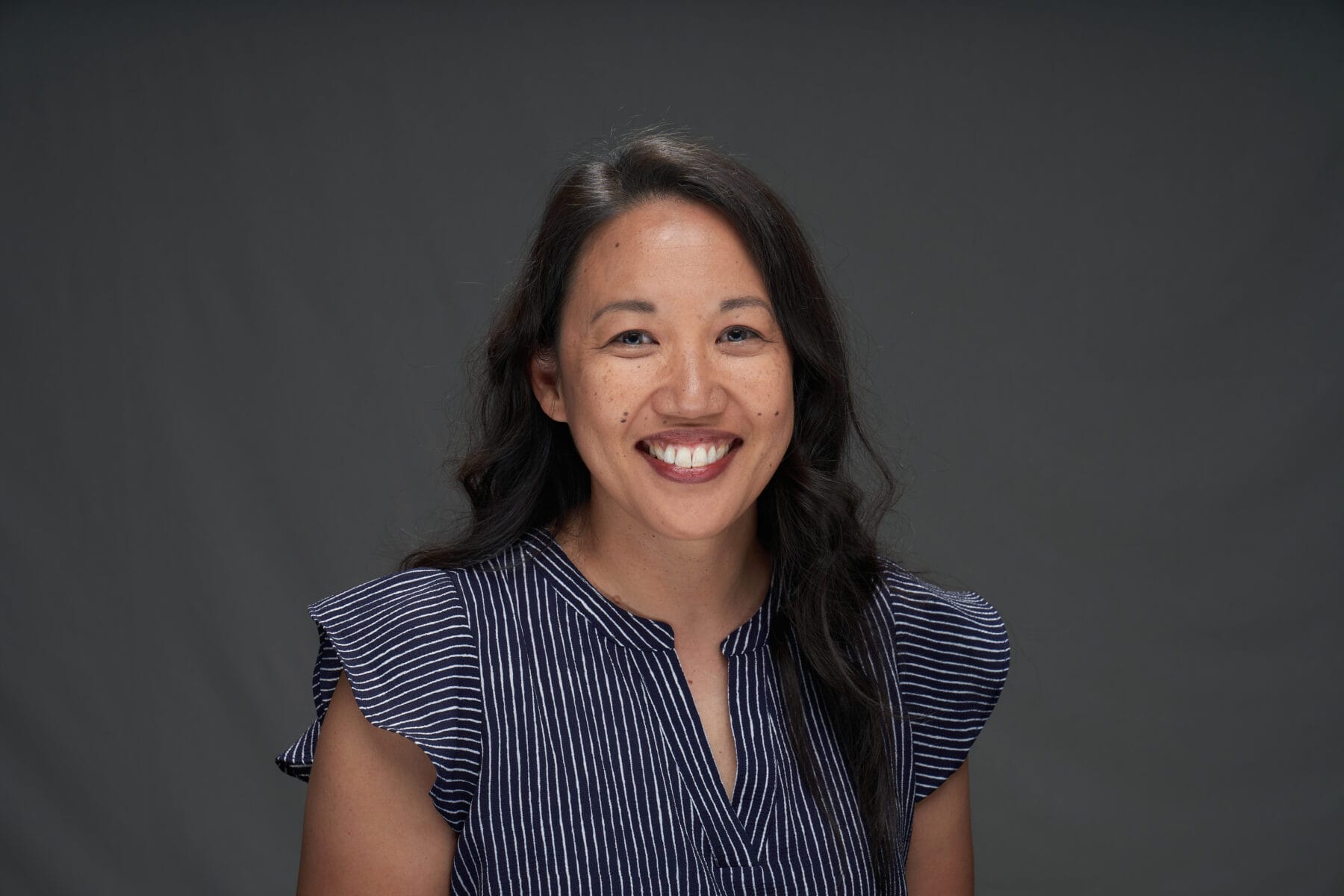 A woman with long dark hair, wearing a navy blue and white striped blouse, smiles at the camera in front of a plain gray background.