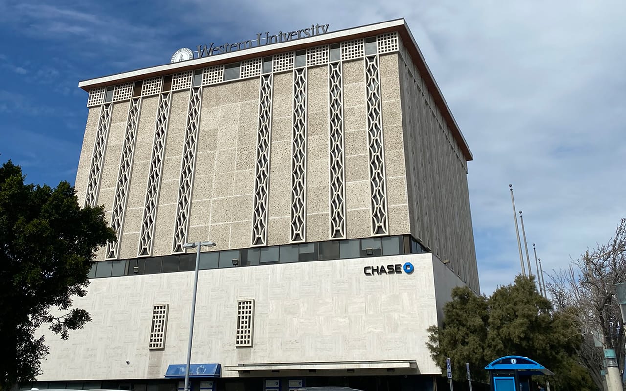 A multi-story building with Western University signage on top. The lower level features a Chase bank office. The facade includes decorative patterns. Trees and a blue structure are visible around the building.
