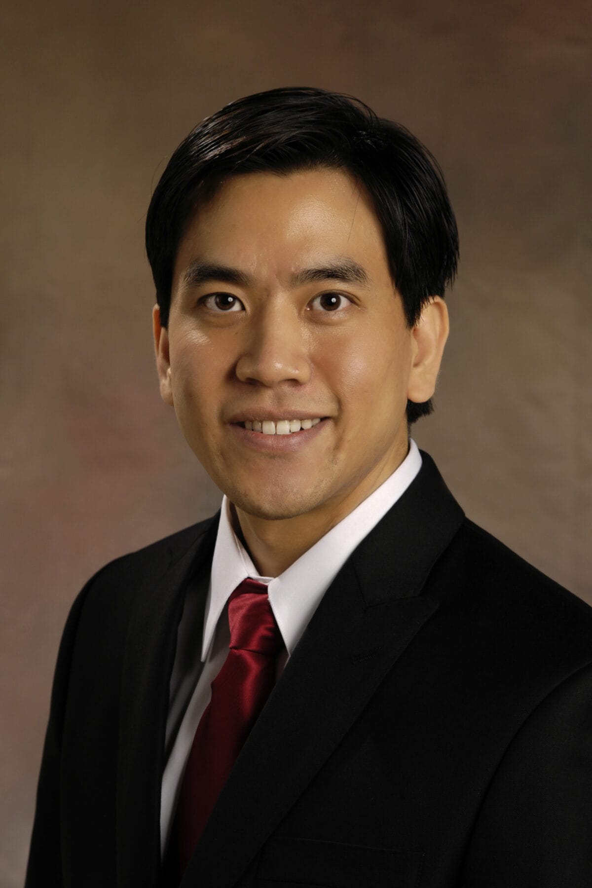 A person wearing a black suit and red tie is smiling at the camera against a neutral background.