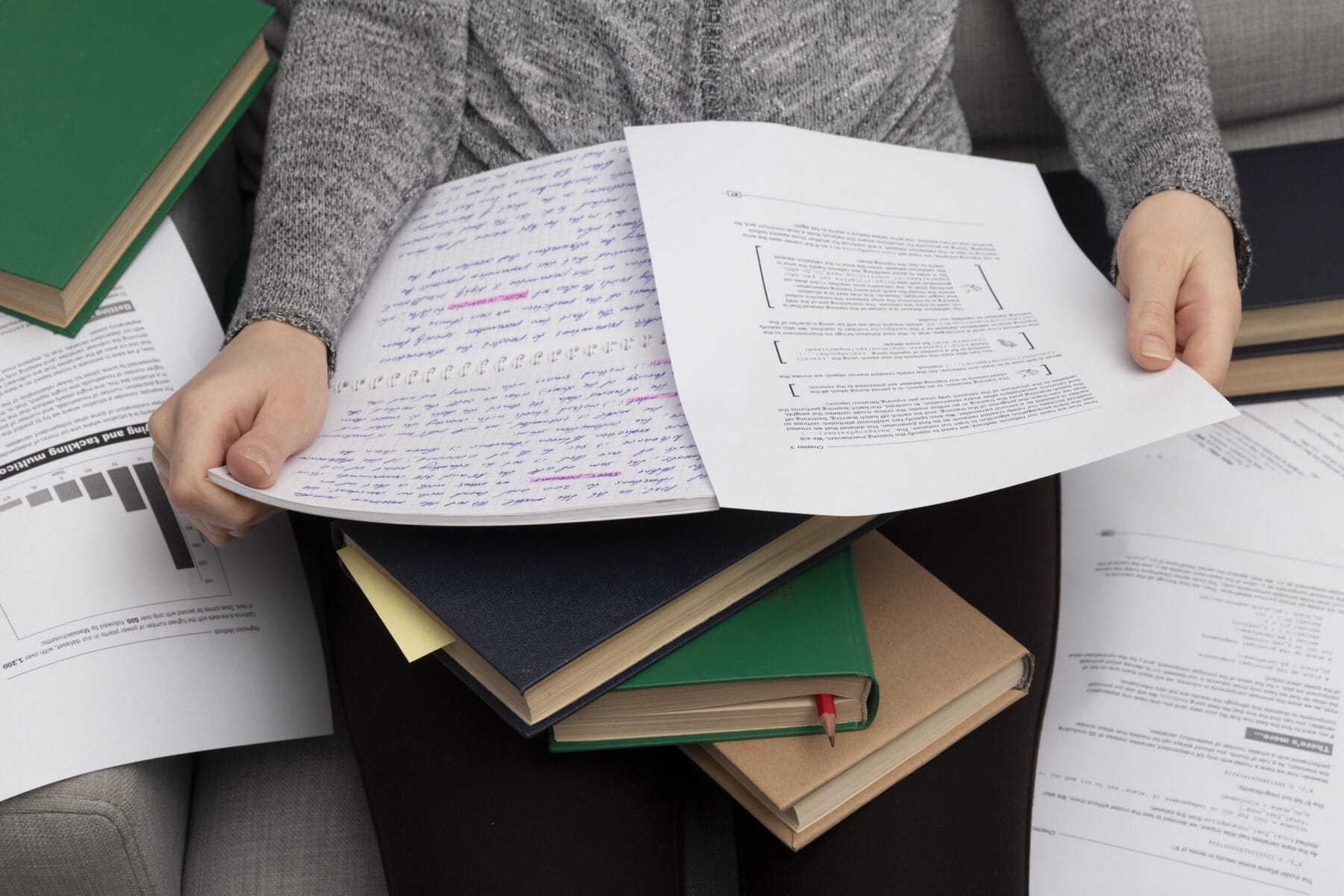 A person sits holding handwritten notes and printed documents, with several books and papers scattered on their lap and around them.