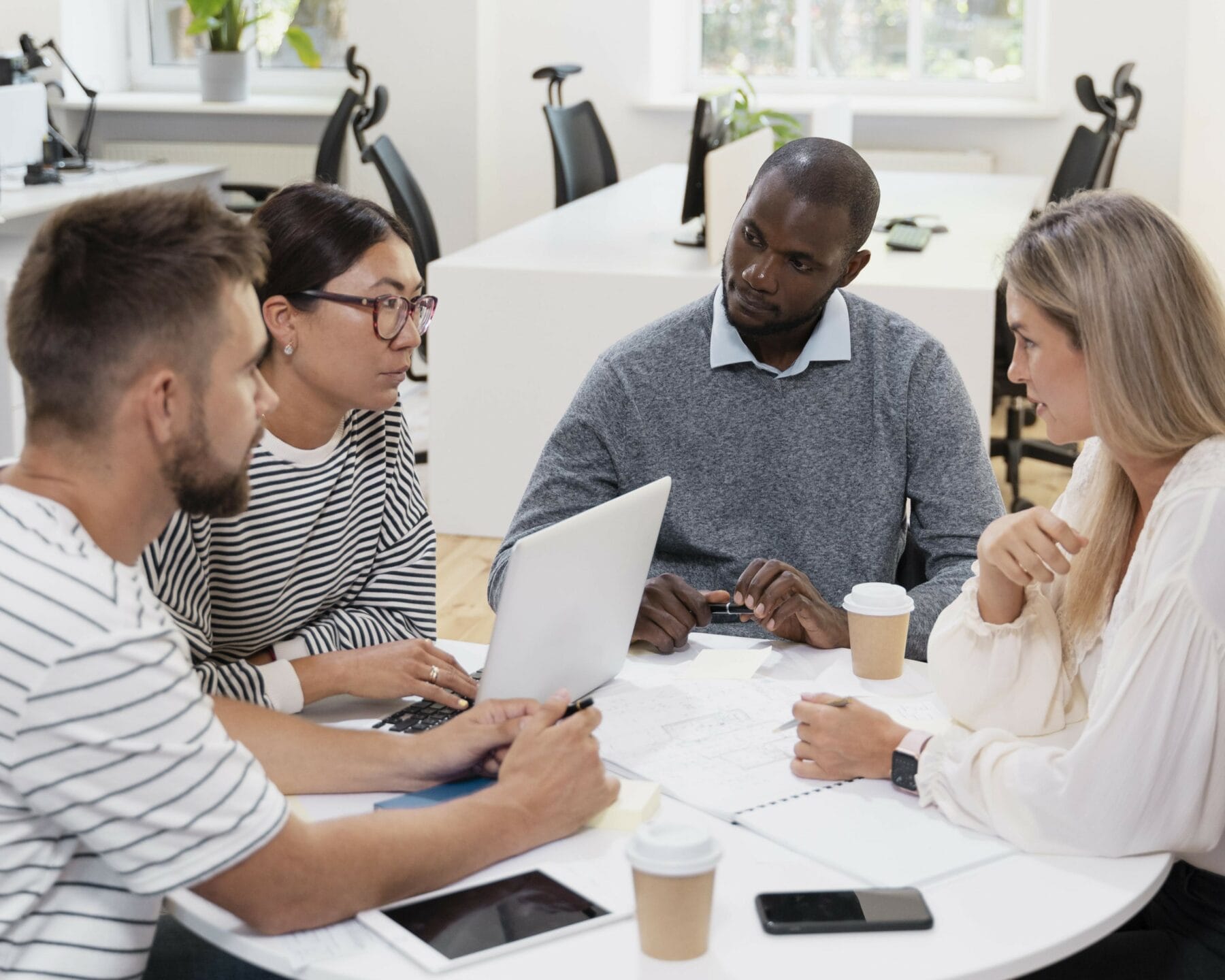 Four people are seated around a table in an office, discussing work. They have notebooks, a laptop, and coffee cups on the table.