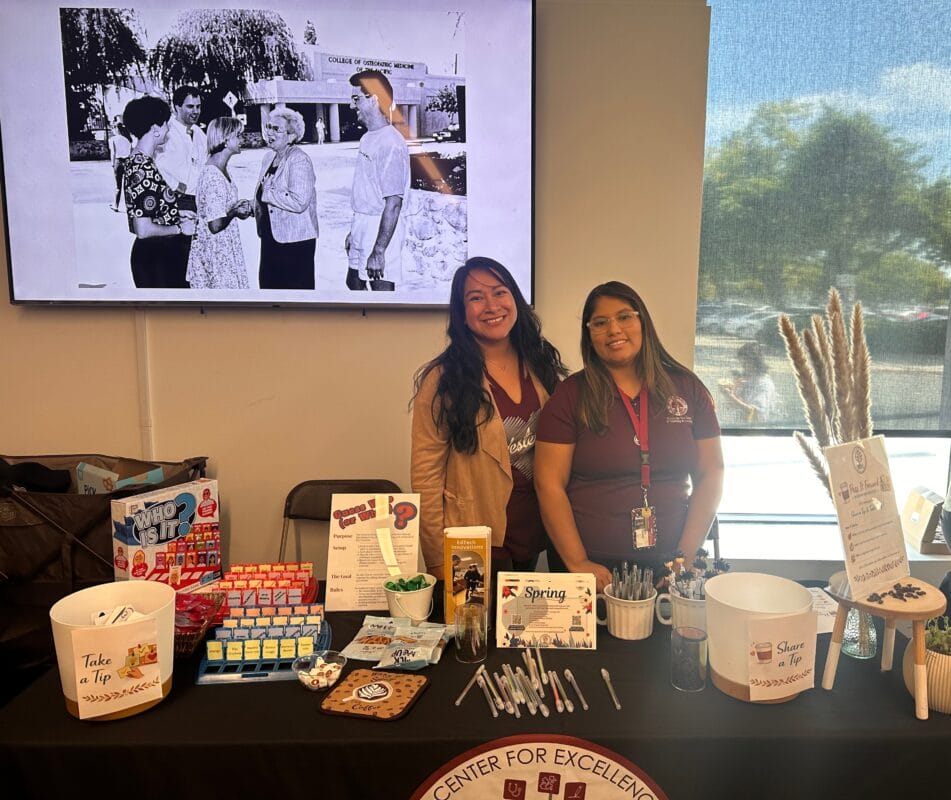 Two women stand behind a table displaying pens, snacks, and informational materials at an indoor event. A black-and-white photo is displayed on the wall behind them.