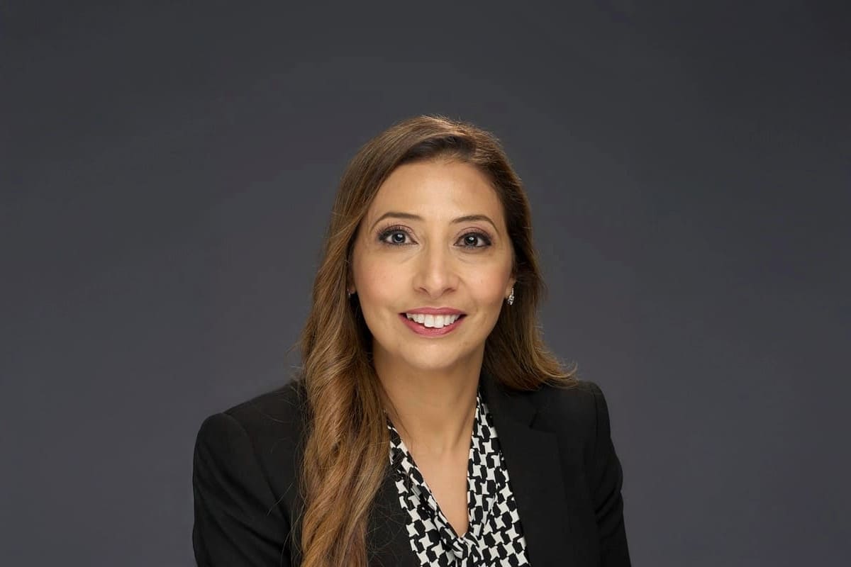 Woman with long brown hair wearing a black blazer and patterned blouse, sitting and smiling in front of a plain dark background.