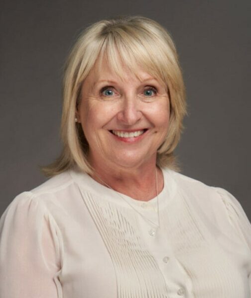 A portrait of a smiling middle-aged woman with blonde hair, wearing a white blouse against a grey background.