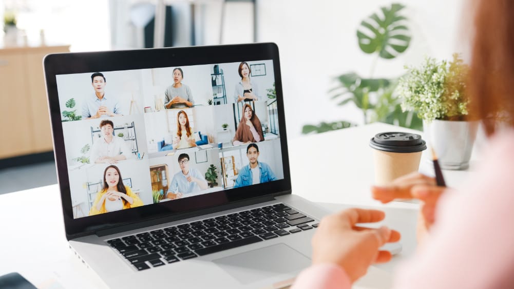 A person is using a laptop to participate in a video conference with eight other individuals displayed in a grid layout on the screen.