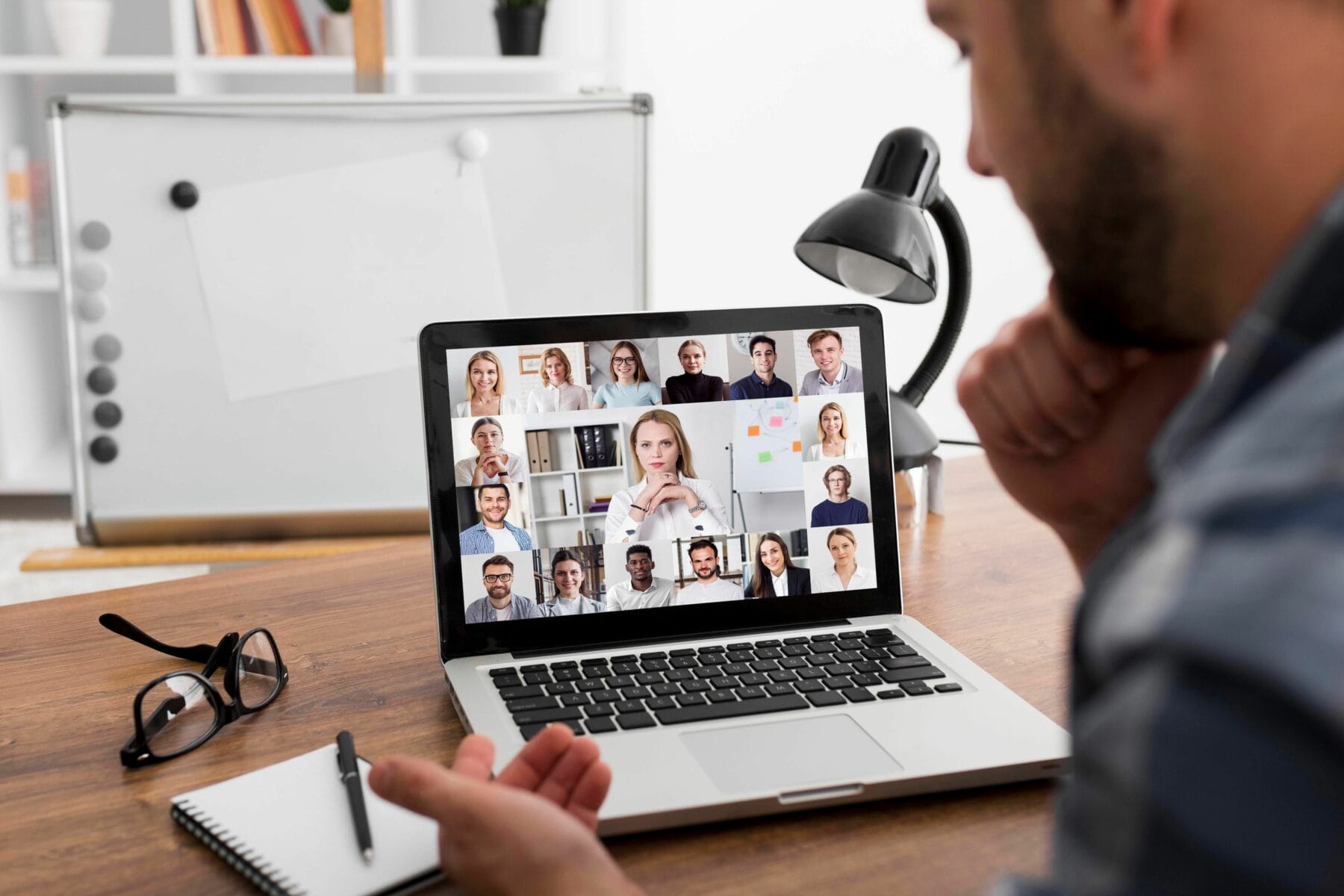 A person participates in a video conference on a laptop, with multiple people visible on the screen. There is a notebook, pen, glasses, and a desk lamp on the wooden table.