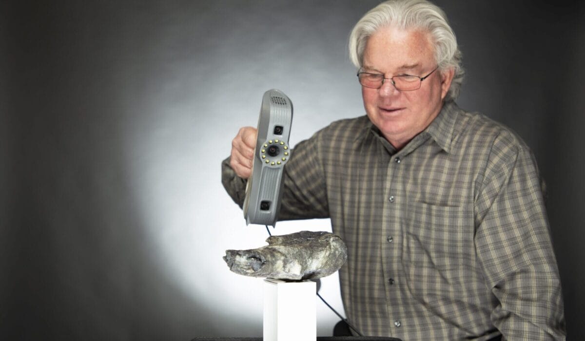 An elderly man in glasses uses a handheld 3D scanner on a fish fossil, with a dark background.