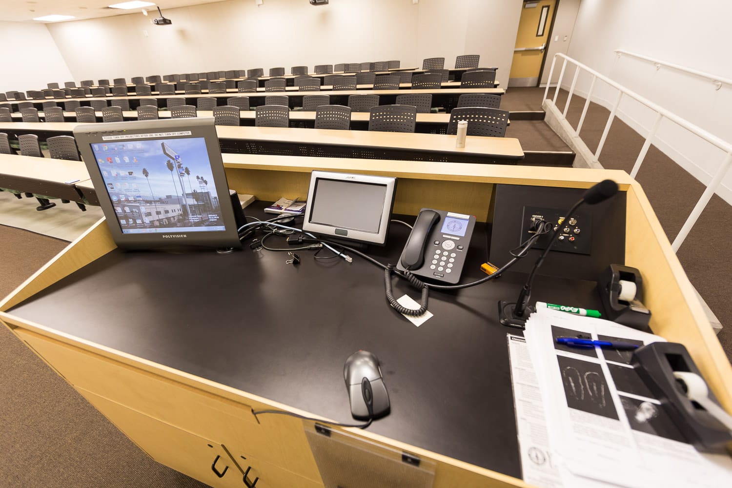 A lecture podium equipped with a computer monitor, tablet, phone, microphone, and various office supplies overlooks an empty classroom with rows of seats.