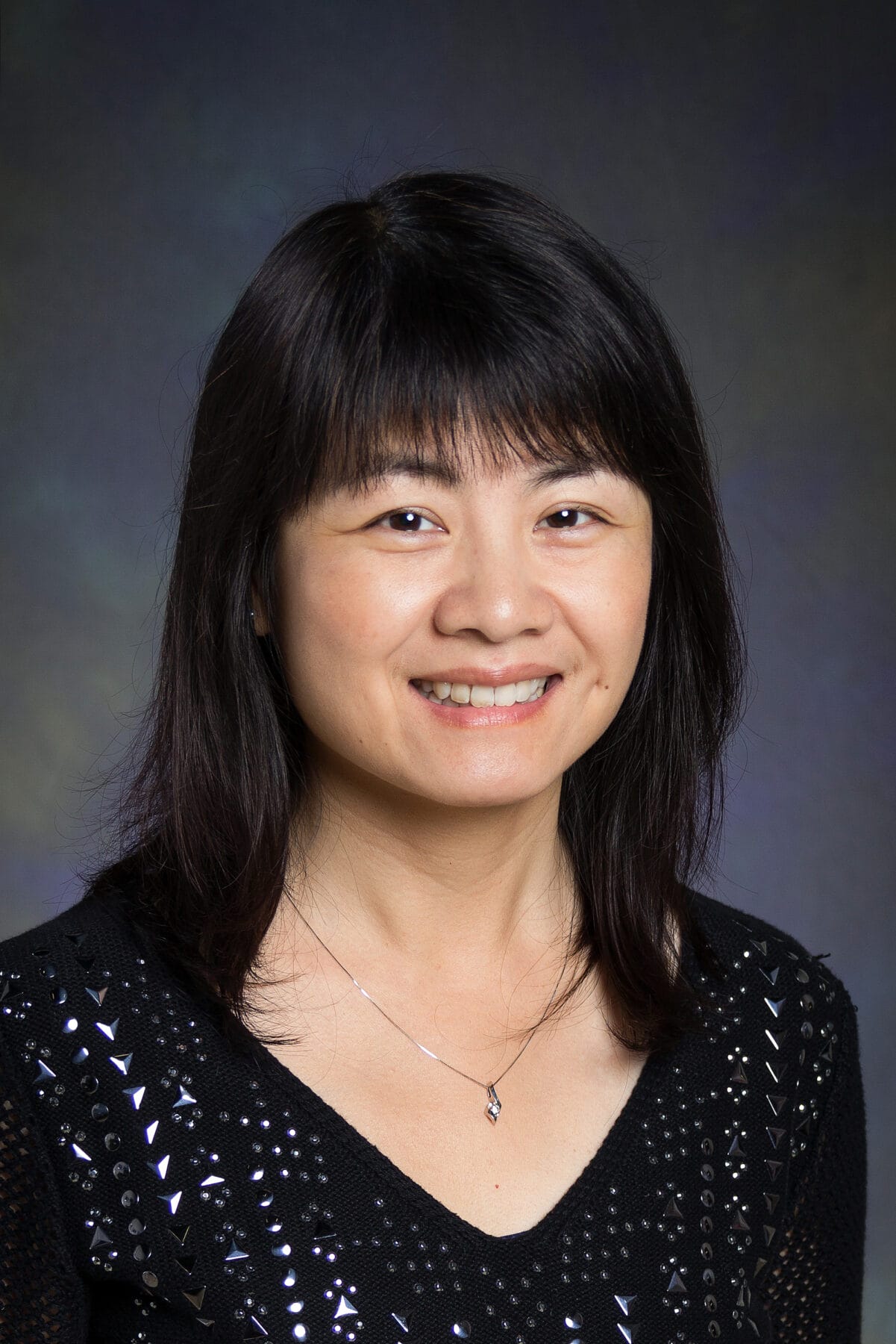 A person with long, dark hair and a black top smiles in a portrait against a dark backdrop.