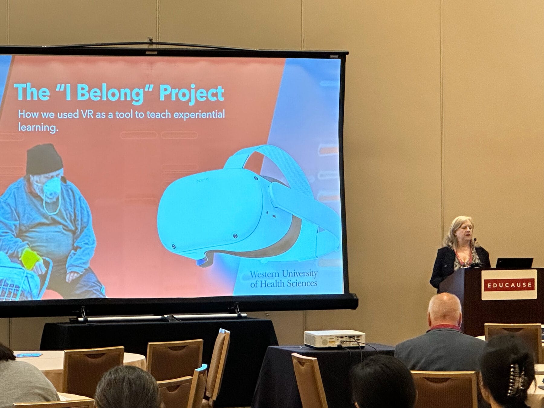 A woman presents a project titled "The 'I Belong' Project" about using VR for experiential learning at a conference. A headset and person in a VR environment are shown on the screen.