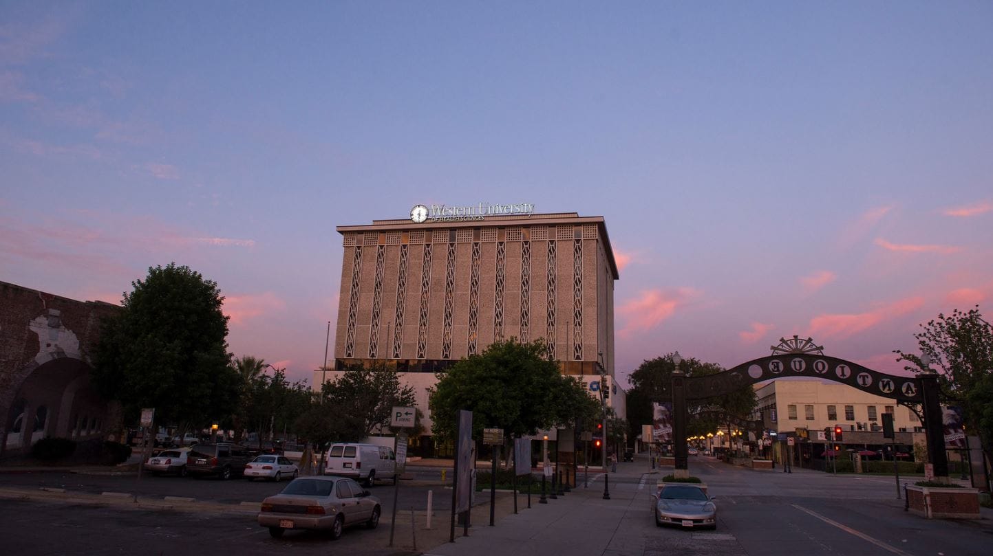A street view at dusk featuring the University of La Verne building, flanked by parked cars and trees. The sky has a gradient of blue and pink hues.