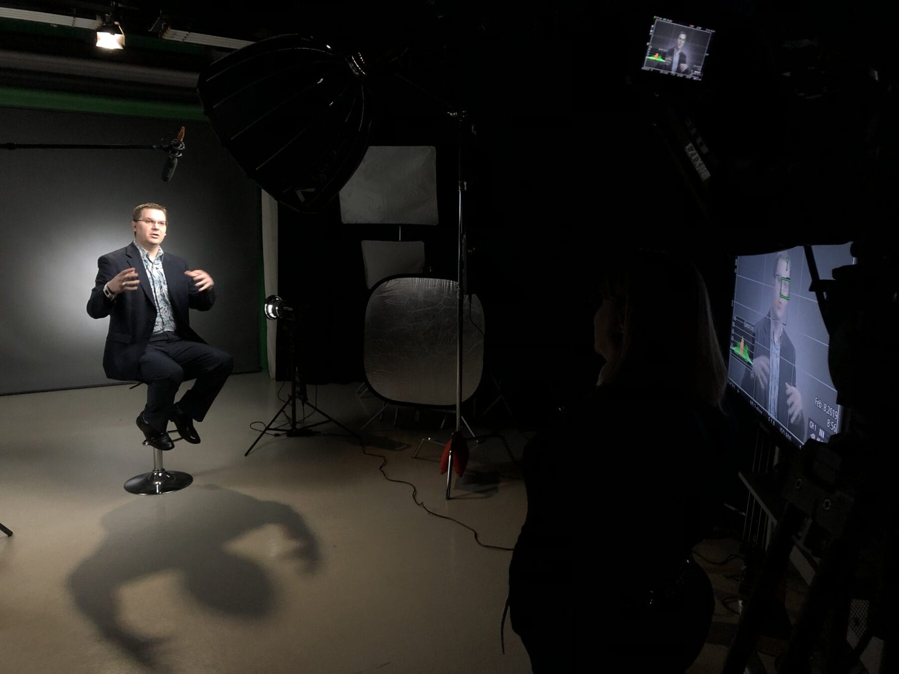 A man in a suit sits on a stool under studio lights being filmed, while two people observe the scene.