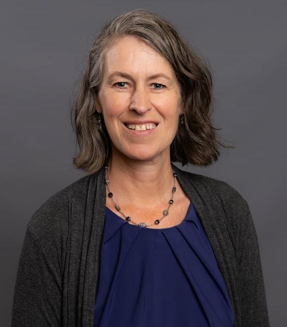 A woman with shoulder-length brown hair, wearing a dark cardigan, blue top, and a necklace, smiles against a gray background.
