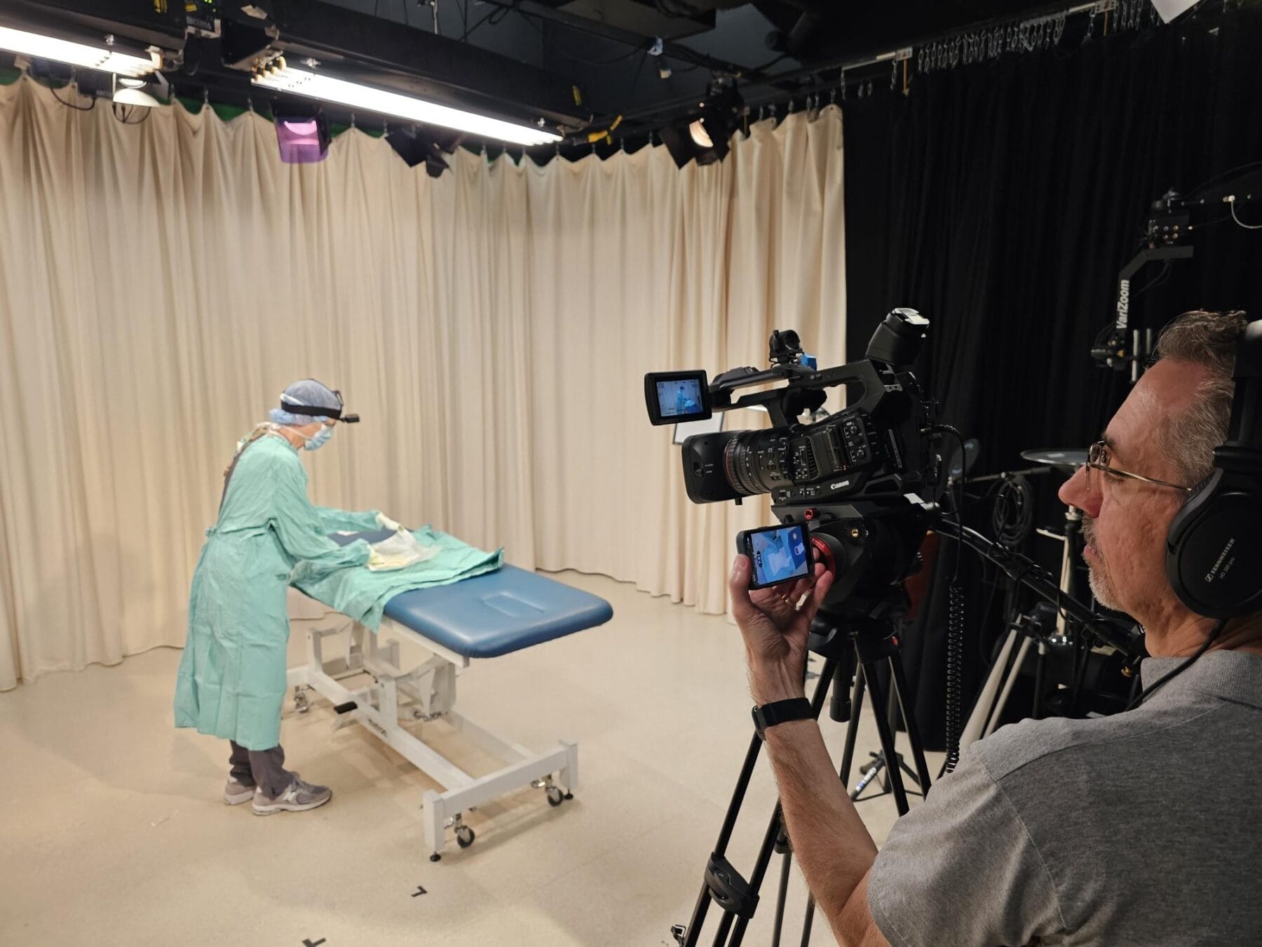 A person in medical scrubs prepares a procedure table while a cameraman films in a studio setting.