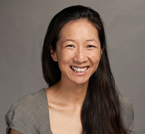 A smiling asian woman with long dark hair wearing a gray blouse, against a neutral gray background.